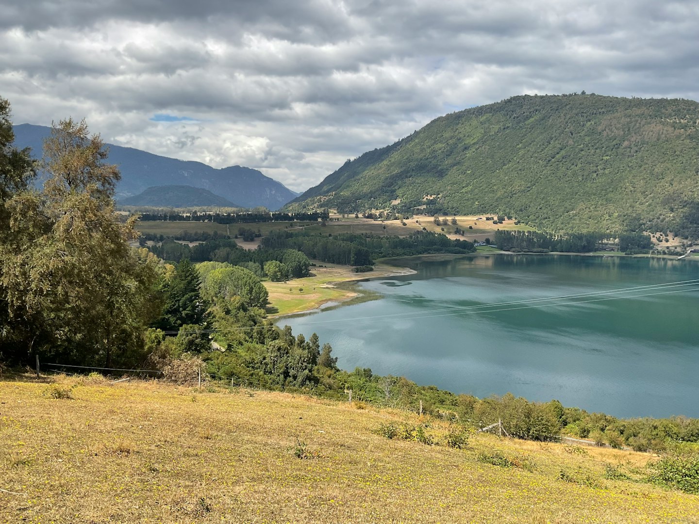 Un lago calmo tra colline boscose e montagne lontane.
